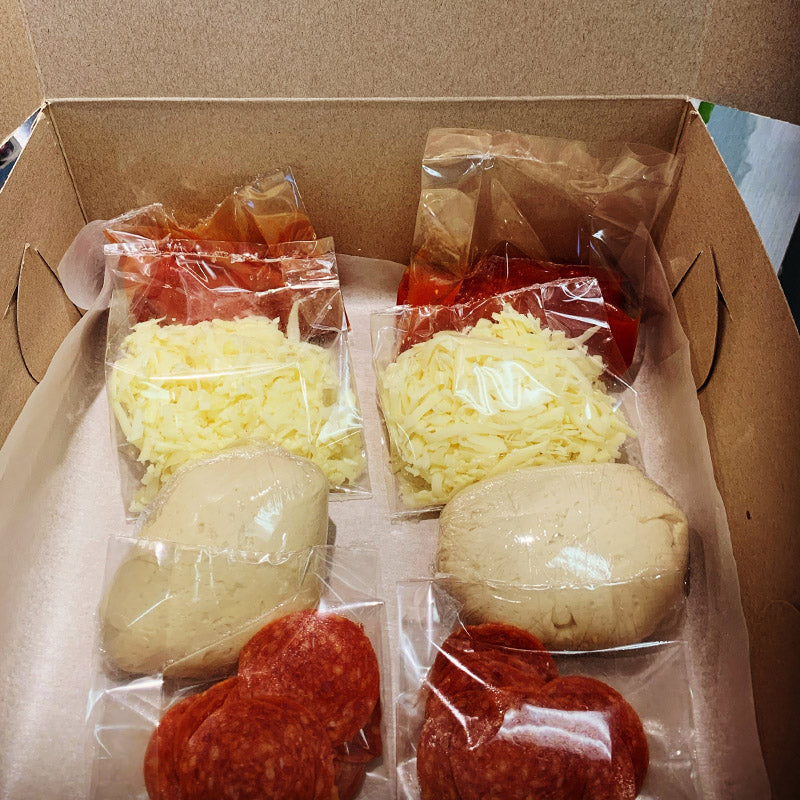 Box filled with pizza ingredients, including two balls of dough, two packages of tomato sauce, and 2 sets of both cheese and pepperoni topping.
